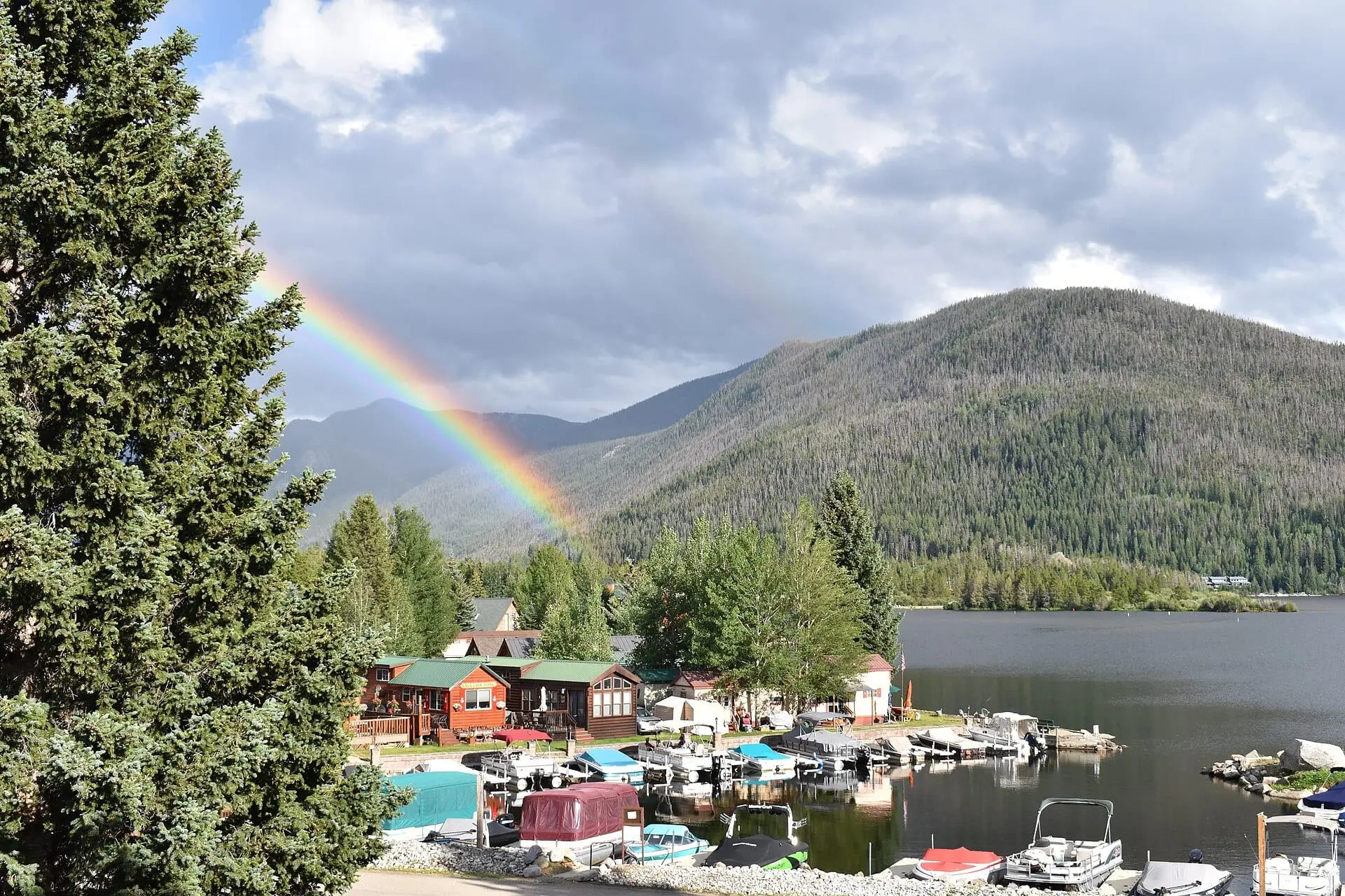 A rainbow over the forested mountains above Grand Lake, with cabins and moored boats along the shore below