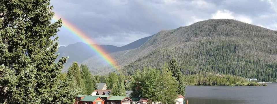 A rainbow over the forested mountains above Grand Lake, with cabins and moored boats along the shore below