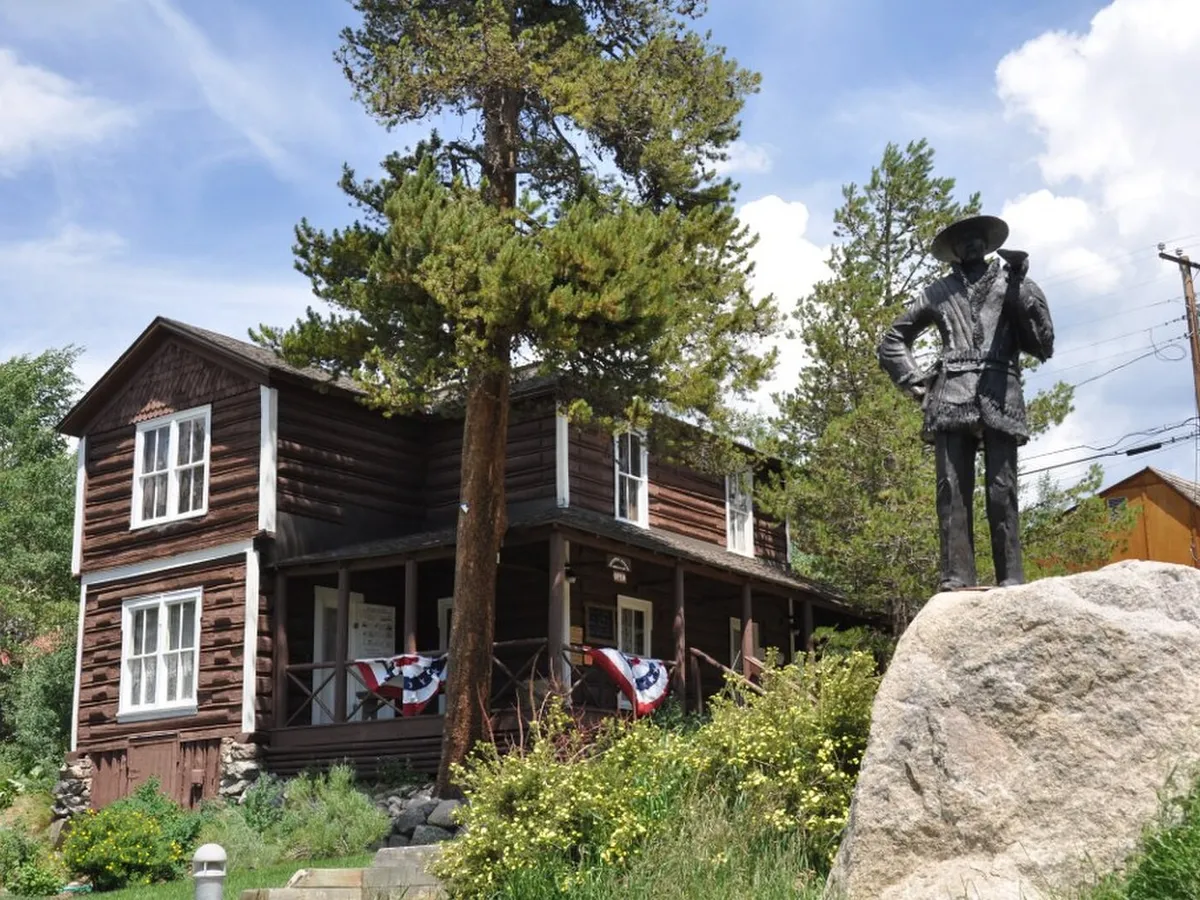 The dark-brown log Kauffman House Museum in Grand Lake, with flag bunting on its porch and a bronze statue on a boulder in front, in 2012