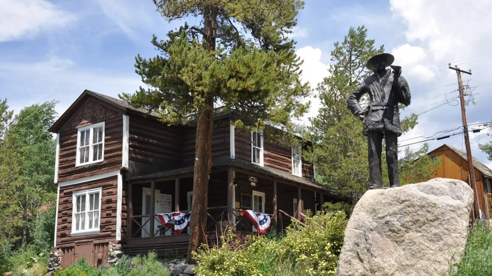 The dark-brown log Kauffman House Museum in Grand Lake, with flag bunting on its porch and a bronze statue on a boulder in front, in 2012