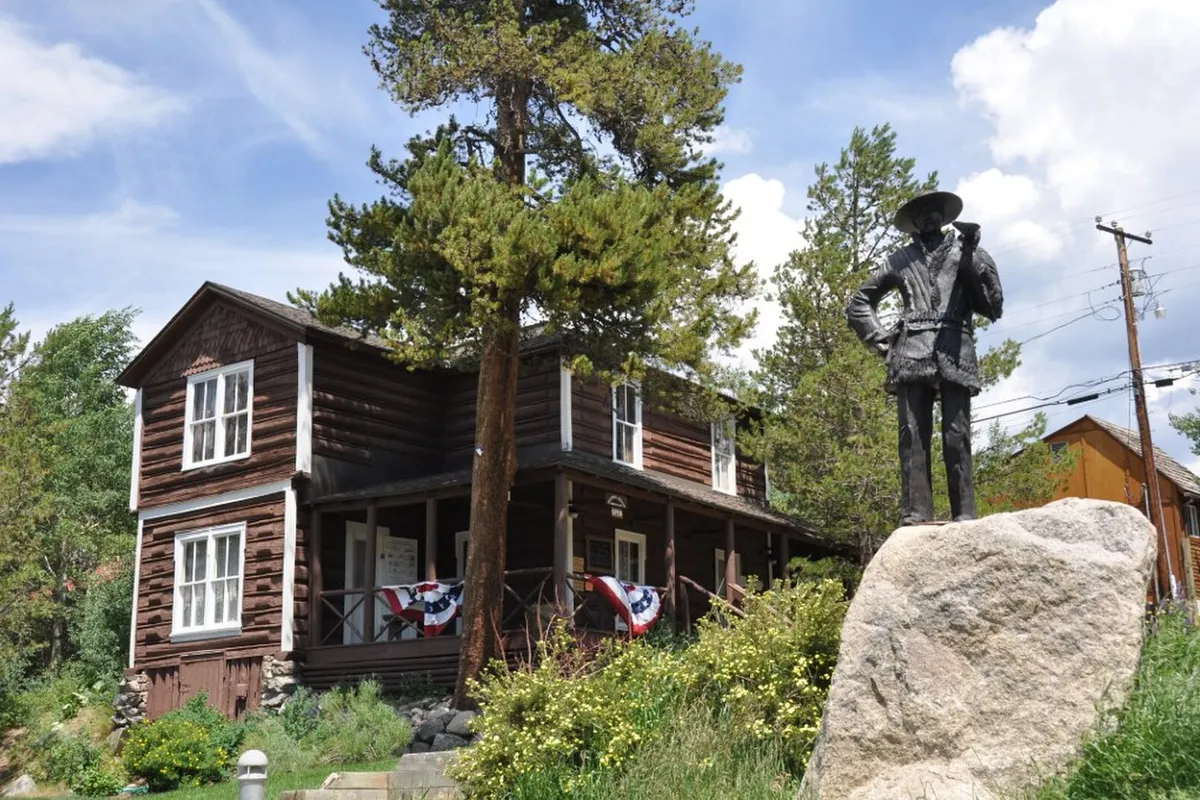 The dark-brown log Kauffman House Museum in Grand Lake, with flag bunting on its porch and a bronze statue on a boulder in front, in 2012