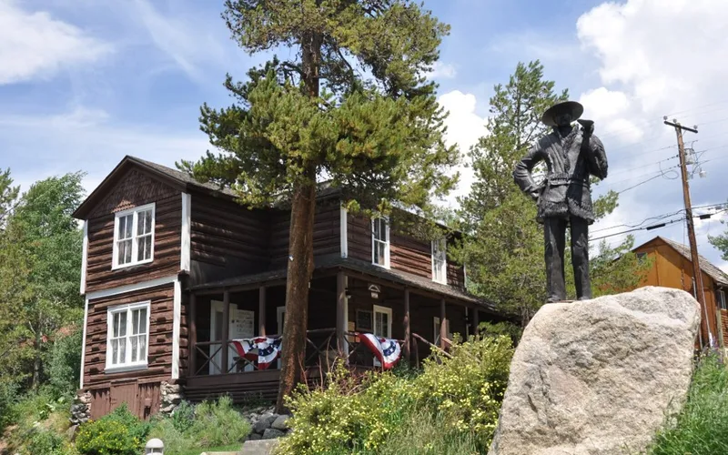 The dark-brown log Kauffman House Museum in Grand Lake, with flag bunting on its porch and a bronze statue on a boulder in front, in 2012